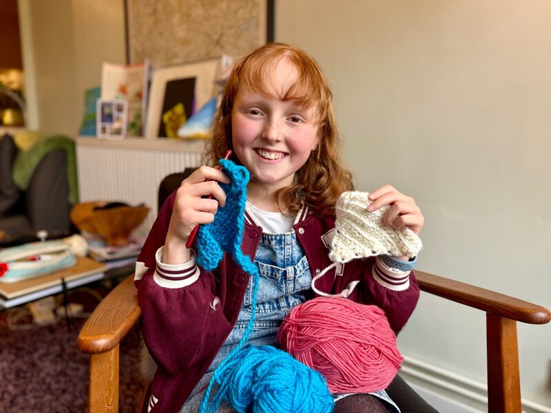 Kirsty smiling, holding crochet bunting triangles in blue and cream, with balls of pink and blue yarn on her lap